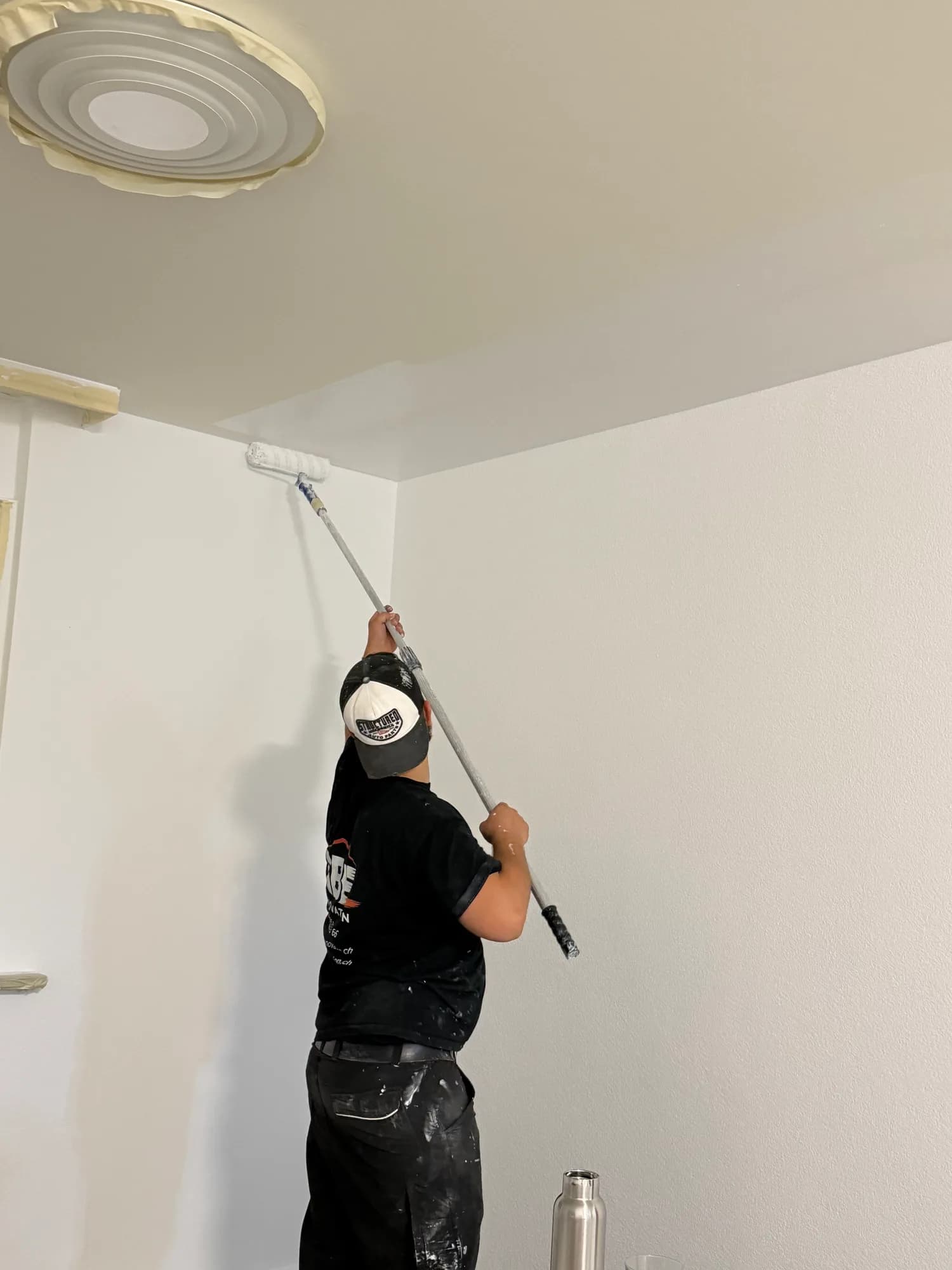 Un ouvrier de dos, t-shirt noir au logo ABE Rénovation, peint un plafond au rouleau à manche, à côté d'une rosace protégée par du ruban de masquage