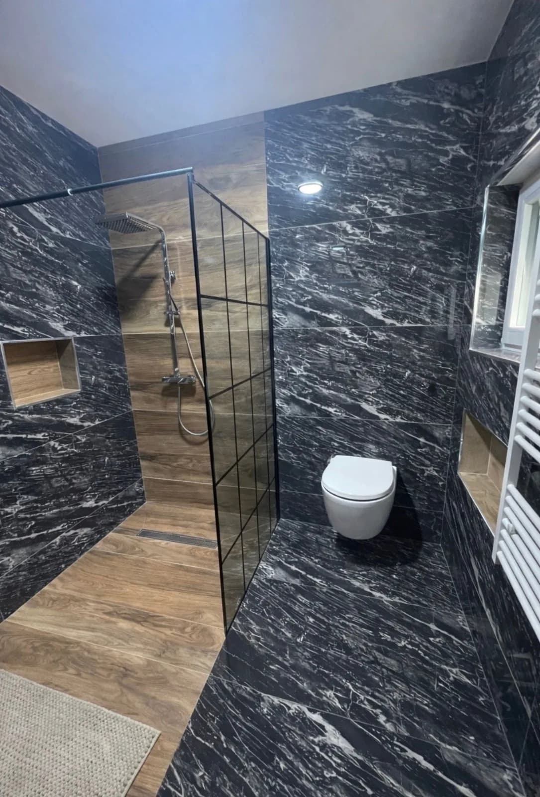 Salle de bains en grands carreaux noirs veinés effet marbre : douche à l'italienne derrière une paroi vitrée, niches encastrées et WC suspendu