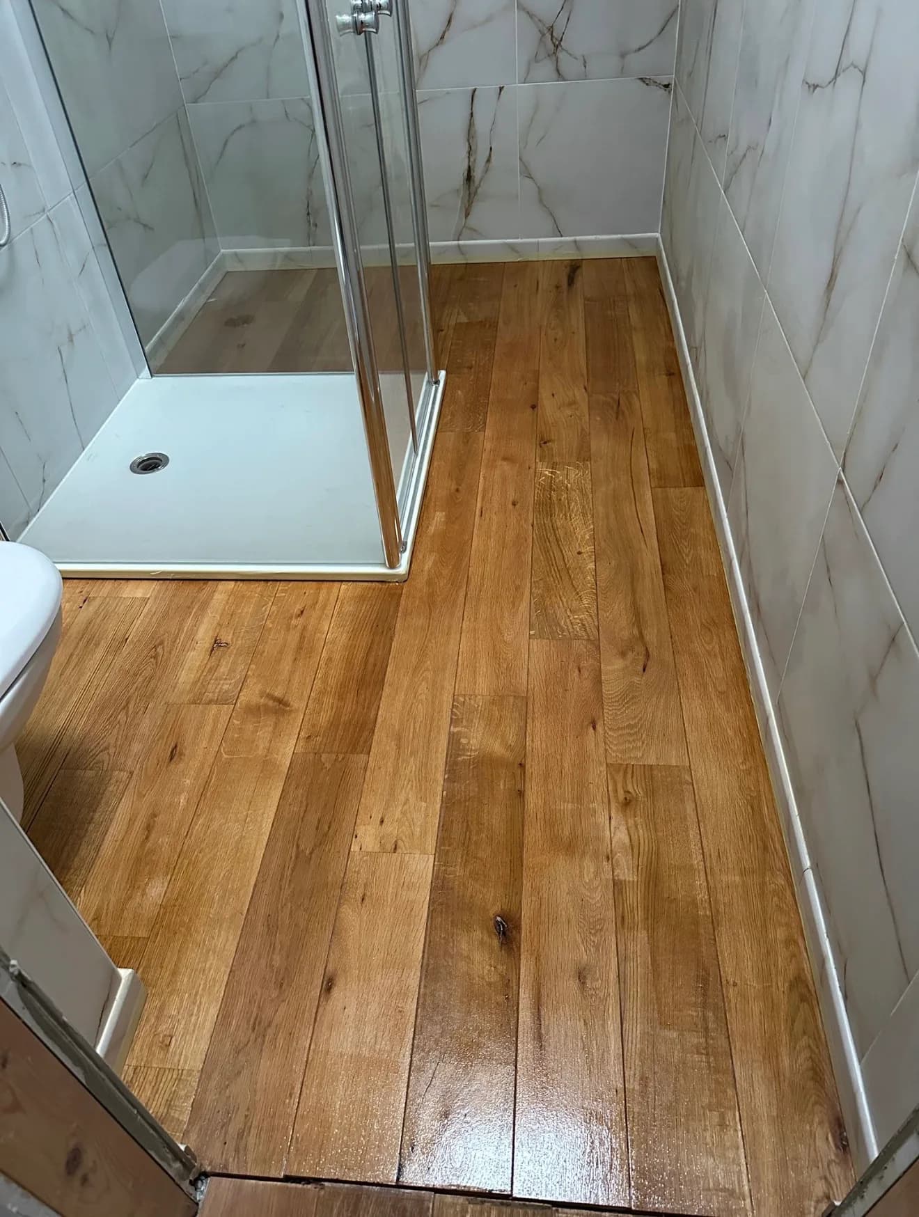 Petite salle d'eau terminée : murs en carreaux blancs effet marbre, douche vitrée et sol en lames d'aspect bois teinte miel