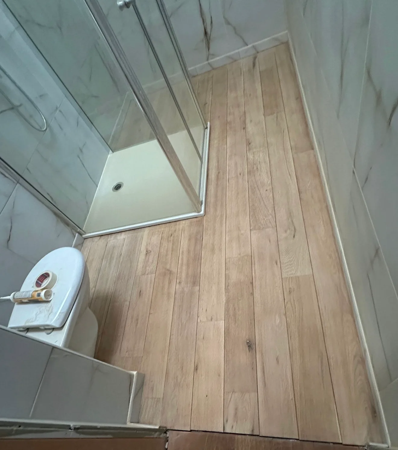 La salle d'eau pendant les travaux, vue d'en haut : sol en lames d'aspect bois clair et mat, cabine de douche vitrée, un rouleau de ruban adhésif et une cartouche de mastic posés sur le WC